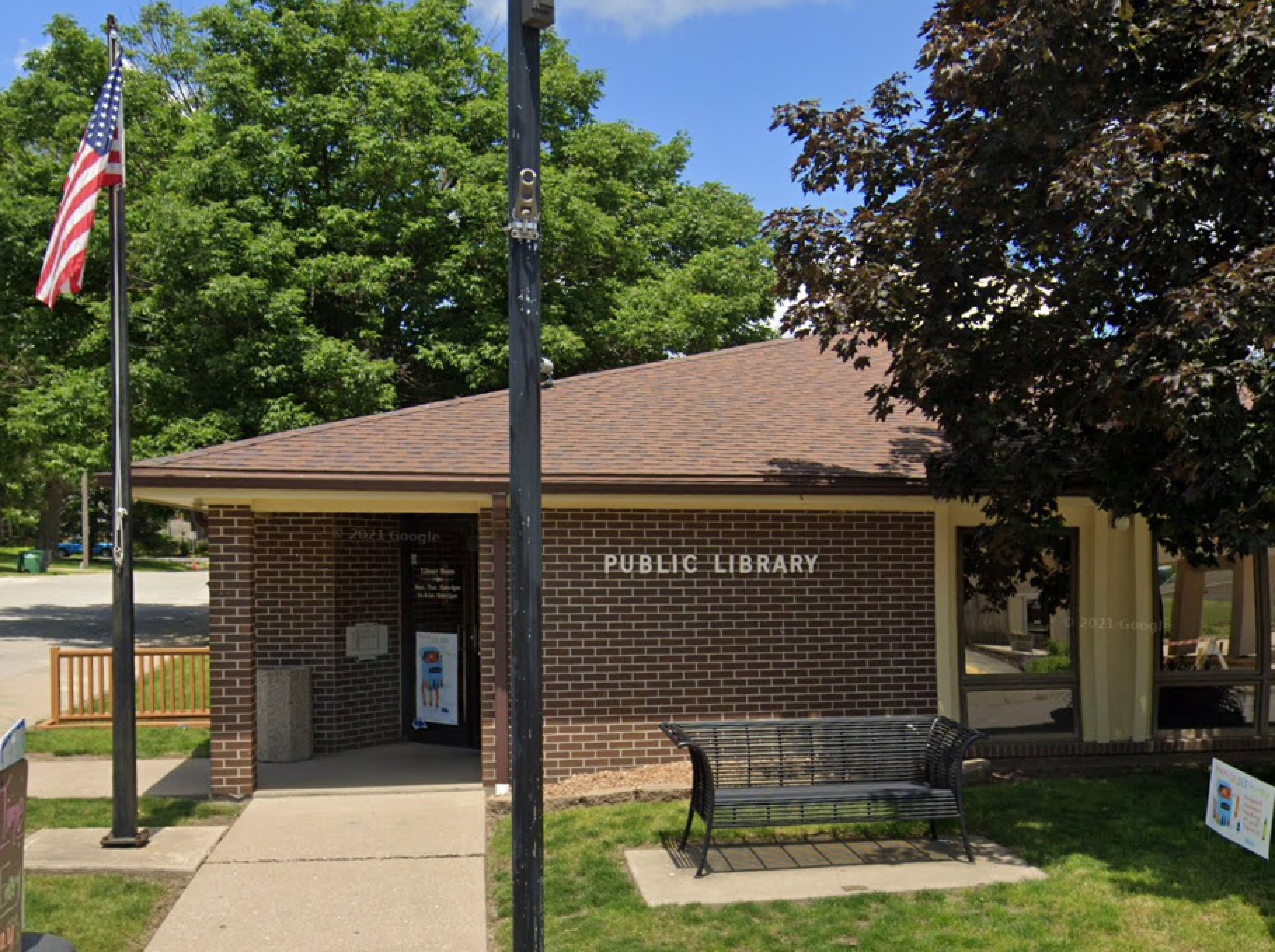 A small public library with a brick exterior and an American flag mounted on a pole in front. A wooden bench is placed near the entrance, and a large tree provides shade. The building is surrounded by grass and a sidewalk leads to the door.