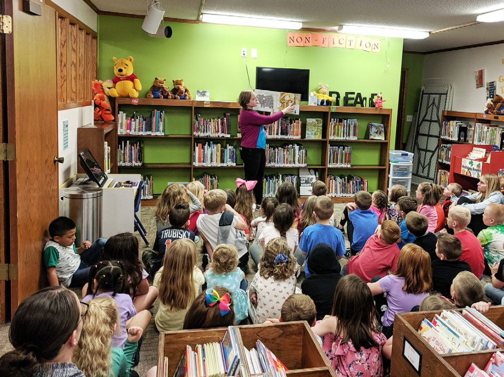 A woman stands reading a book to a large group of children seated on the floor in a vibrant and colorful library. Shelves filled with books and stuffed animals are visible in the background, and a sign reading "NON-FICTION" is displayed above the shelves.