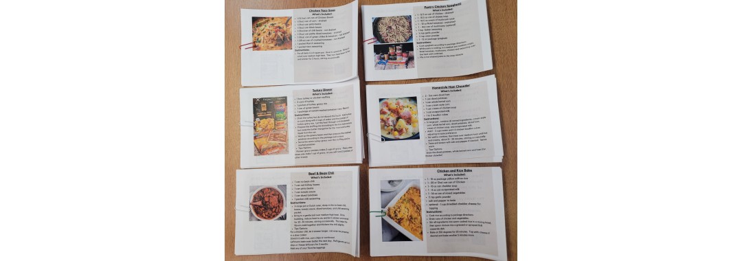 Seven recipe cards with images of dishes and lists of ingredients and instructions are arranged on a wooden table in two rows. Each card features a different meal, with photos and text visible but not readable.